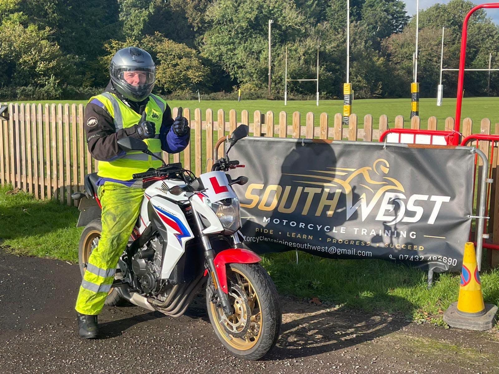 A rider giving a thumbs up on the white Honda CB650F with an L plate, beside the South West Motorcycle Training banner on the training pad at North Tawton A rider giving a thumbs up on the white Honda CB650F with an L plate, beside the South West Motorcycle Training banner on the training pad at North Tawton
