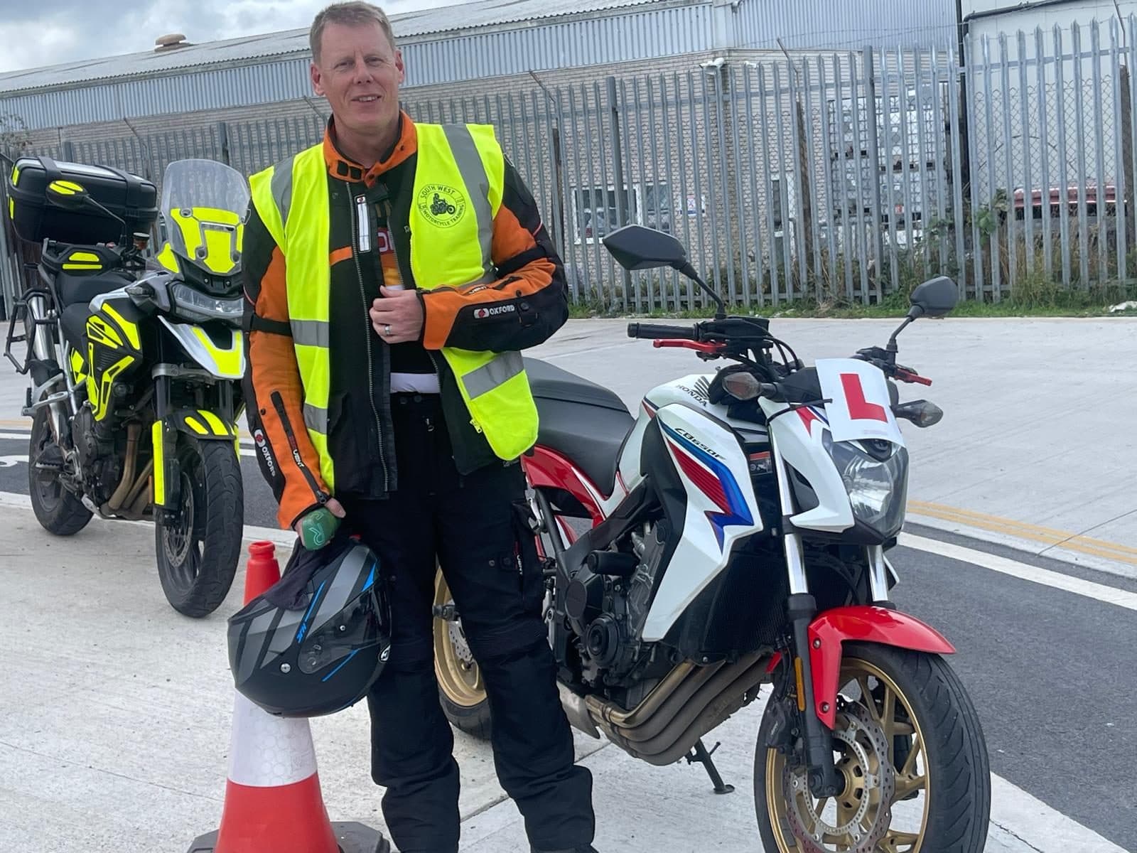 A rider in hi-vis holding his helmet beside the white Honda CB650F with an L plate at the test centre after Module 2 A rider in hi-vis holding his helmet beside the white Honda CB650F with an L plate at the test centre after Module 2