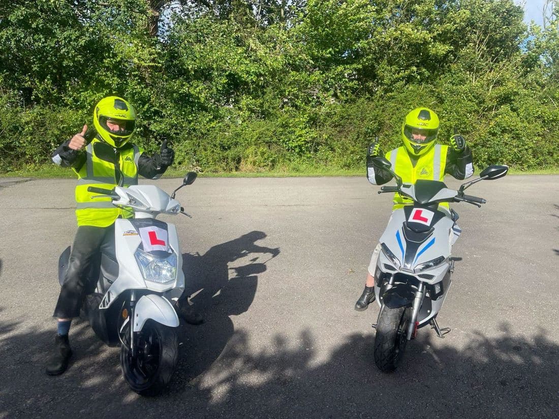 Two learner riders in hi-vis giving a thumbs up on white L-plated mopeds during CBT motorcycle training Two learner riders in hi-vis giving a thumbs up on white L-plated mopeds during CBT motorcycle training
