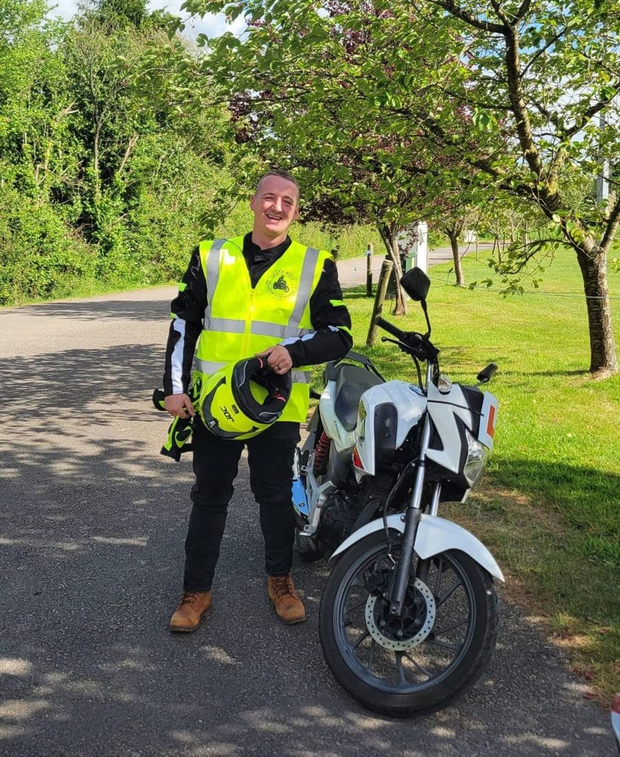 A rider in a South West Motorcycle Training hi-vis holding his helmet beside a white 125cc training motorcycle A rider in a South West Motorcycle Training hi-vis holding his helmet beside a white 125cc training motorcycle