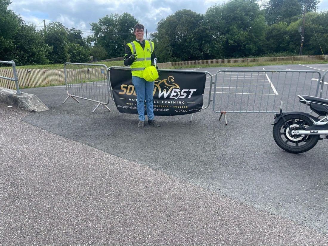 A learner rider in hi-vis with a yellow helmet in front of the South West Motorcycle Training banner on the training pad A learner rider in hi-vis with a yellow helmet in front of the South West Motorcycle Training banner on the training pad