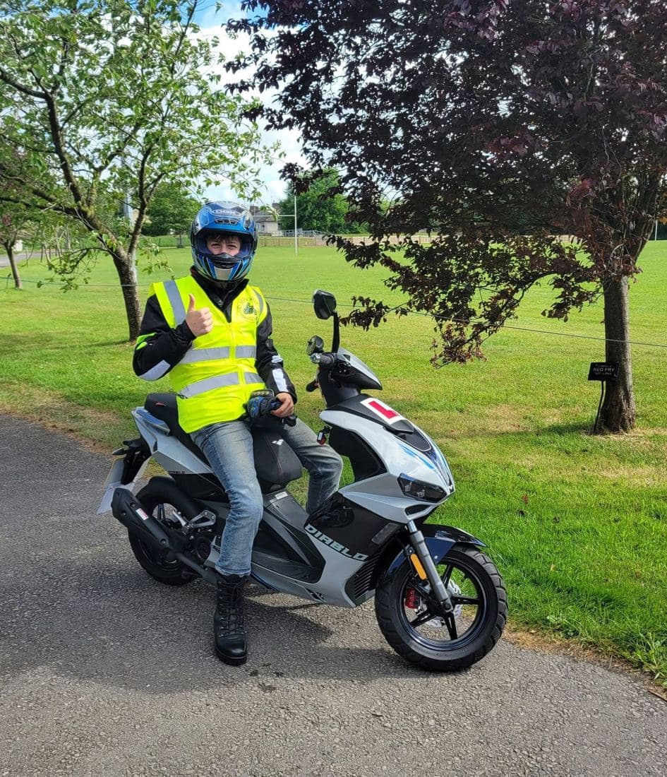 A young learner rider in hi-vis and a blue helmet sitting on an L-plated scooter, giving a thumbs up
